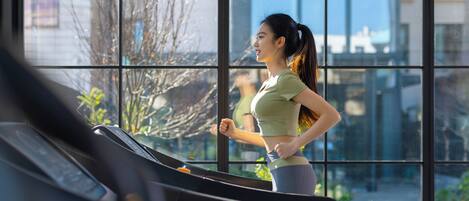 A woman running on a treadmill in a gym with large windows and a view of buildings and trees outside.