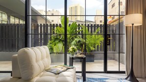 A modern living room with a white sofa, a small side table with a vase of flowers, and a large window with a view of buildings and a fence. - Kunming Kasion Hotel (Kunming)