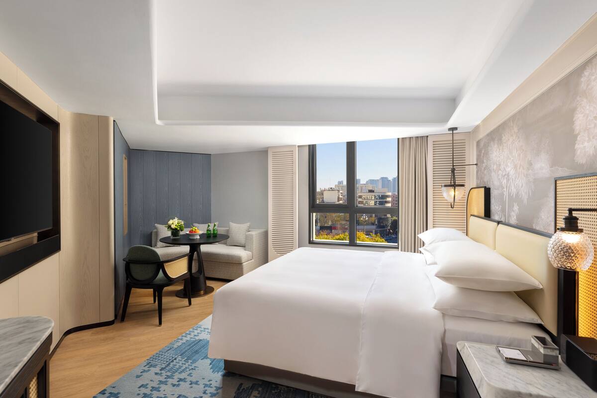A modern hotel room with a large bed, a seating area with a table and chairs, a TV, and a city view through the window.