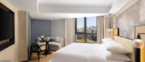 Una habitación de hotel moderna con una cama grande, un área de descanso con una mesa y sillas, un televisor y vistas a la ciudad desde la ventana.