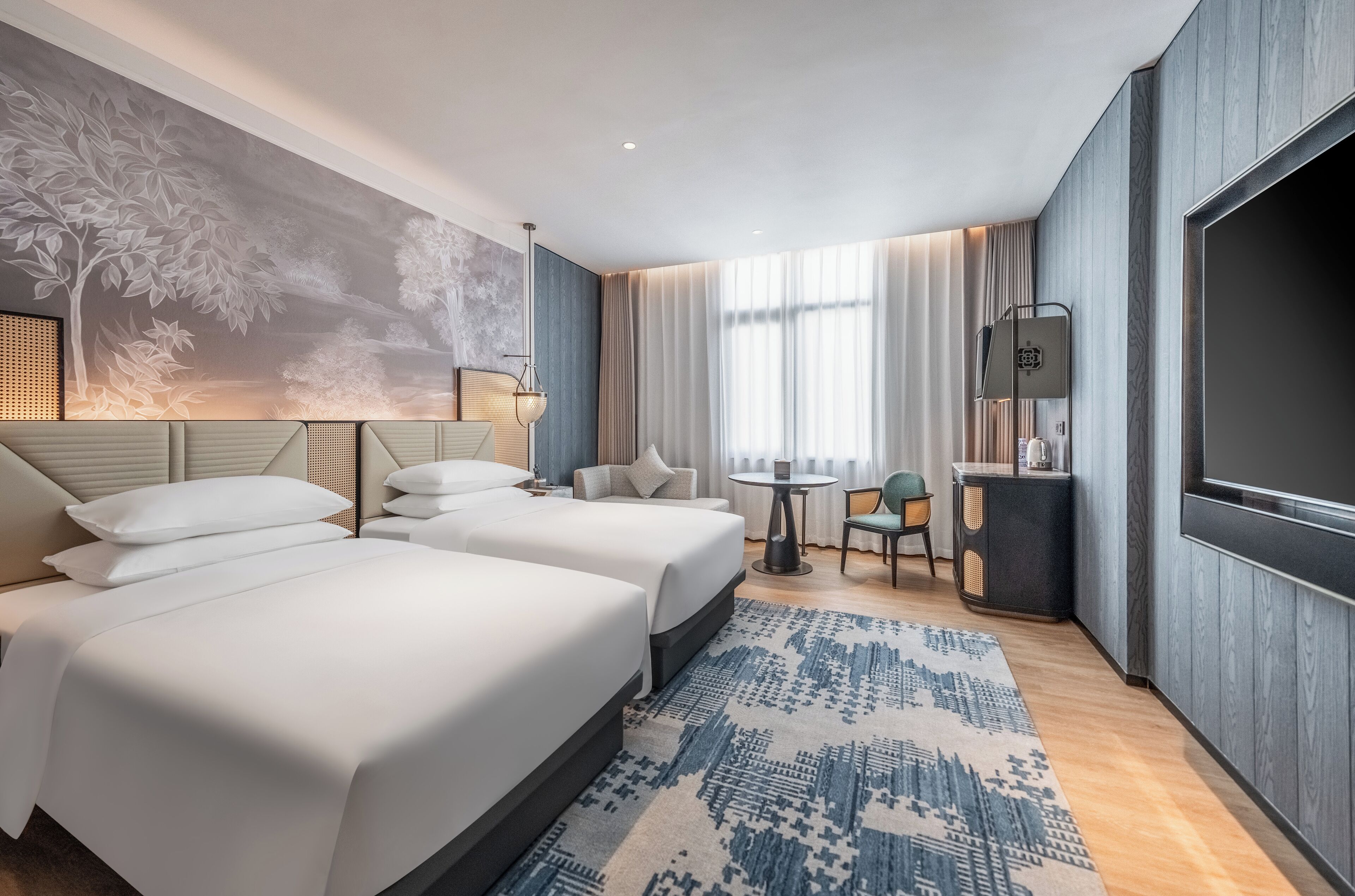 Habitación de hotel con dos camas, un escritorio, una silla, un televisor, un mural en la pared y una alfombra estampada.