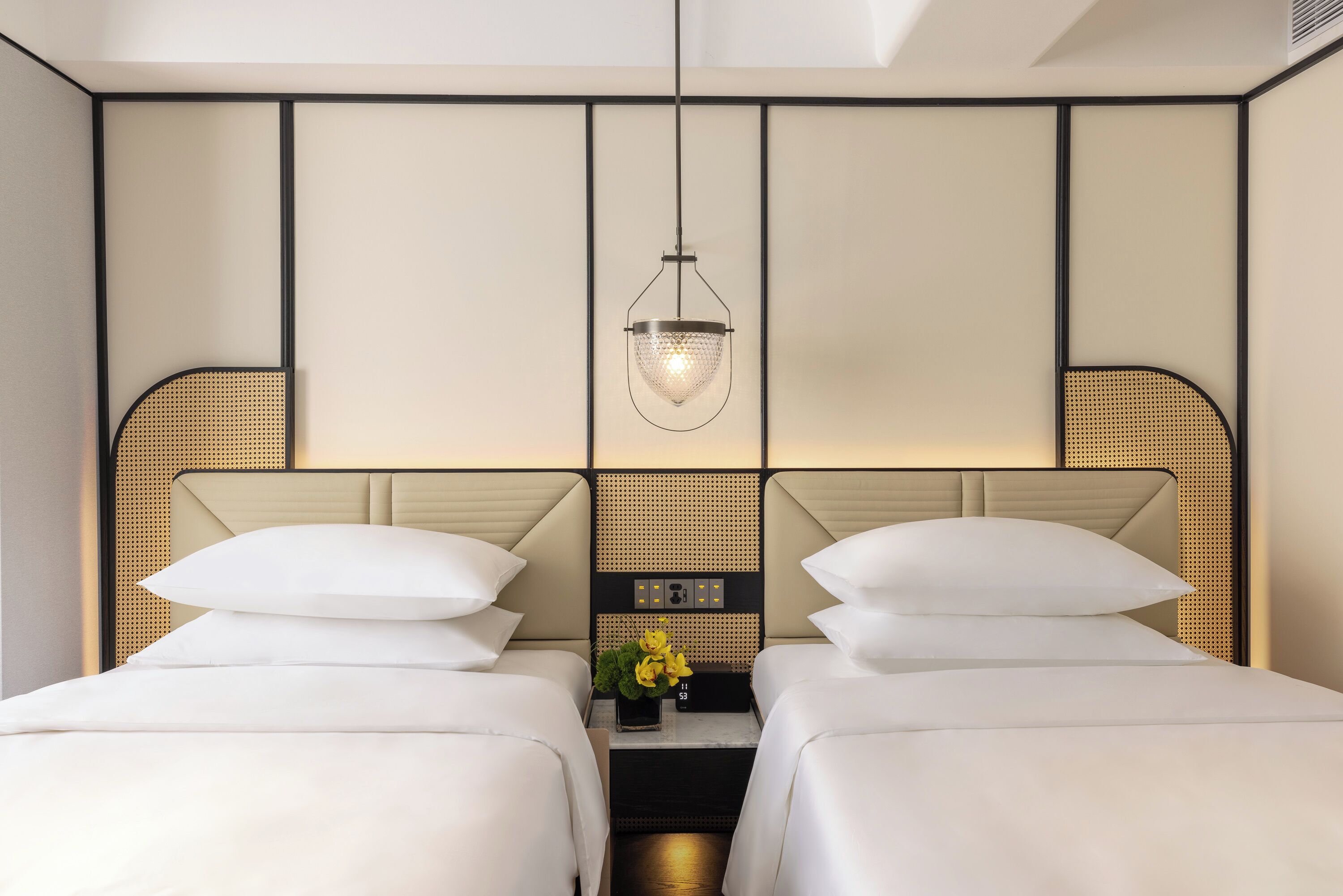 A hotel room with two beds, a nightstand with a clock and a vase of flowers, and a hanging light fixture.