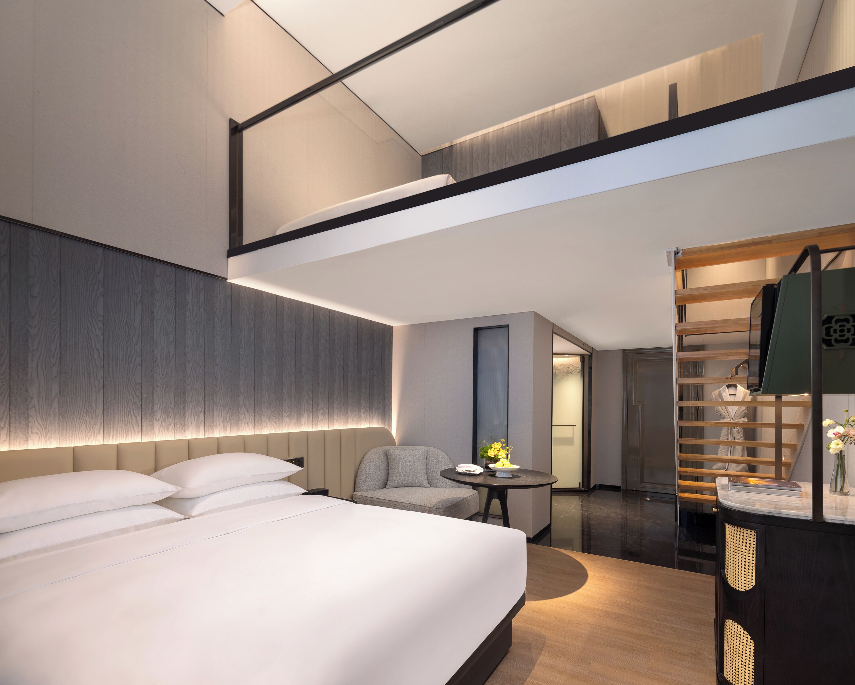 A modern hotel room with a large bed, a seating area, a small table, and a wardrobe.
