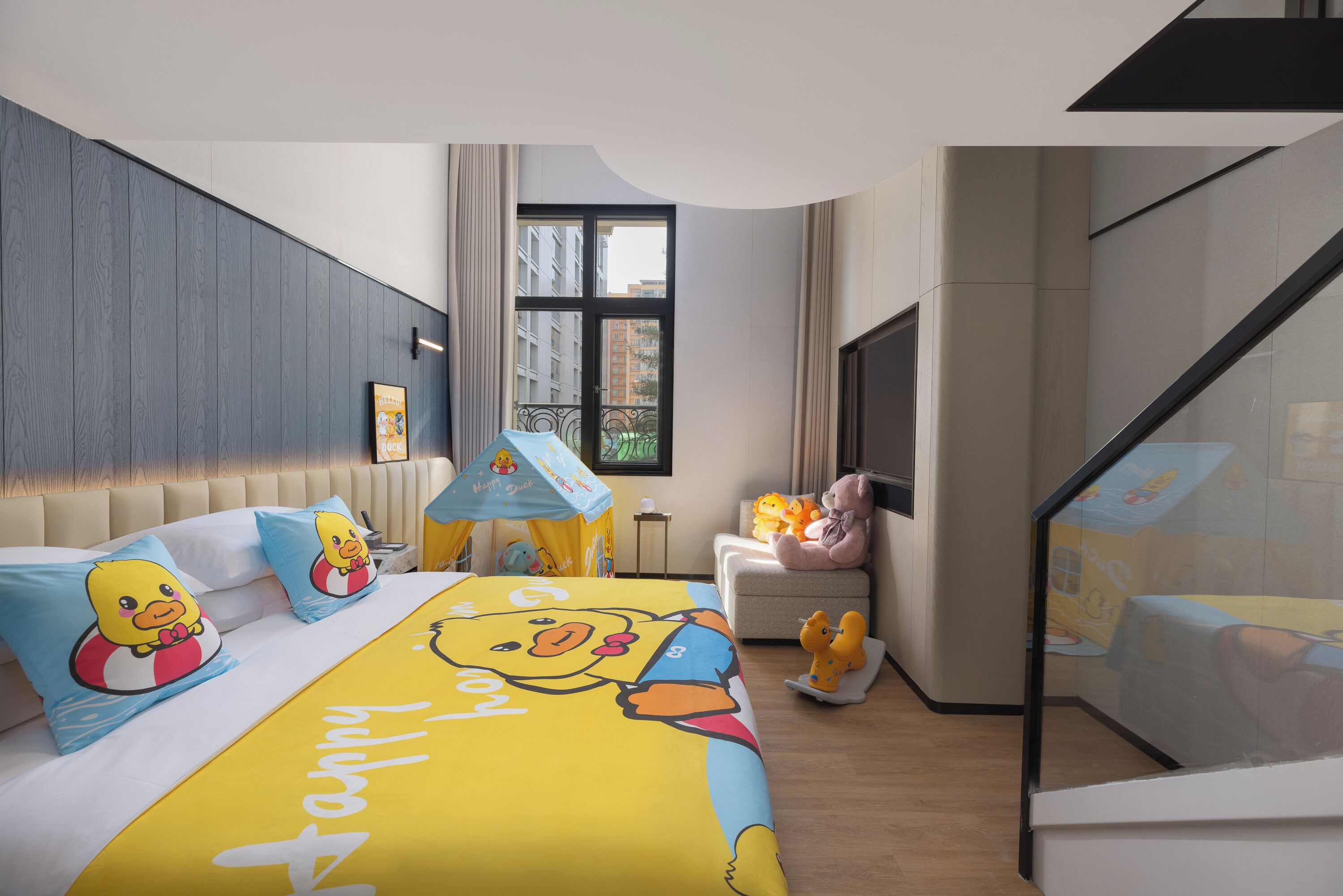Una habitación de hotel moderna con una cama grande, almohadas con forma de pato de dibujos animados, una mesita con lámpara y un balcón con vistas a la ciudad.