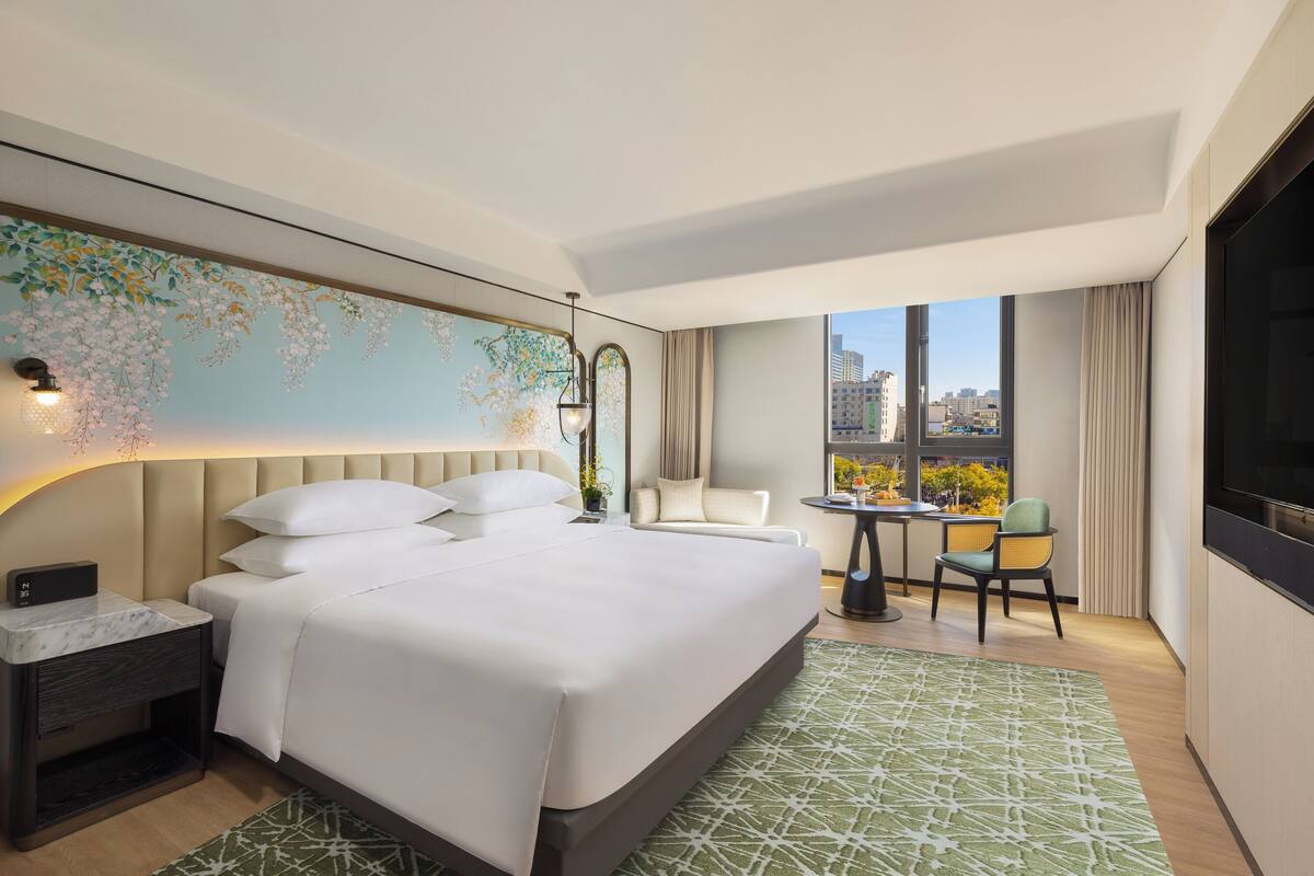 A modern hotel room with a large bed, a desk, and a chair. There is a view of the city through the window.