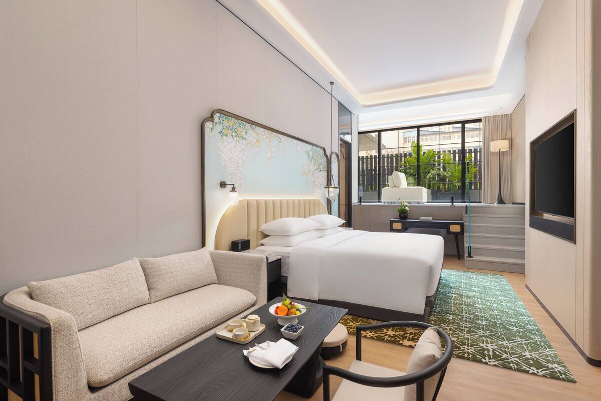 A modern hotel room with a sofa, a dining table, a bed, and a view of the outdoors.