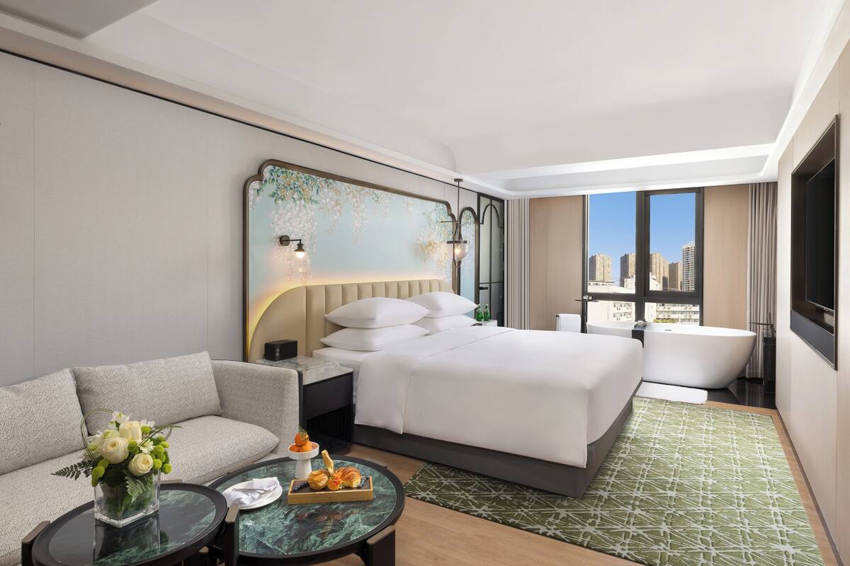 A modern hotel room with a large bed, a sofa, a dining table, and a city view.