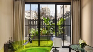 A modern bathroom with a large glass bathtub, a small table with a vase of flowers, and a view of a courtyard with plants and a fence. - Kunming Kasion Hotel (Kunming)