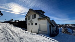 A white building labeled "Alpine Haven" with a sloped roof, situated on a snowy slope with a clear sky and a visible contrail. - Alpine Haven 6 - Mt Hotham (Hotham Heights)