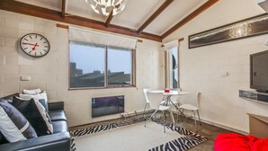 A modern living room with a sofa, a TV, a dining table with chairs, a clock, and large windows. - Alpine Haven 6 - Mt Hotham (Hotham Heights)