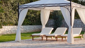 Um gazebo com cortinas brancas, três cadeiras de descanso e um muro de pedra ao fundo.