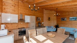 A cozy cabin interior with wooden walls, a dining area with a table and chairs, a kitchenette, a sofa, and a chandelier. - Villa Lara by Villas Guide (Kamenmost)