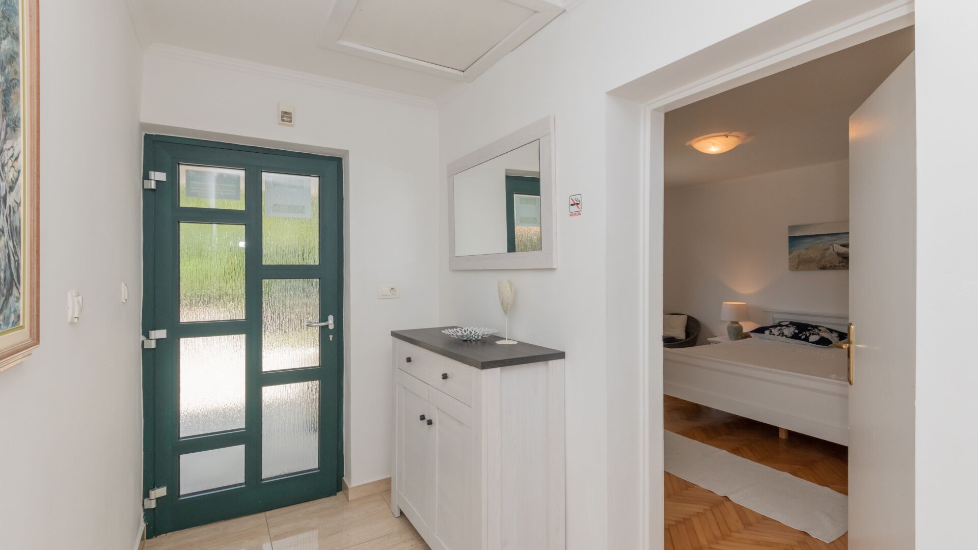 A hallway with a door leading to a bedroom. The room has a bed, a bedside table, and a mirror on the wall.