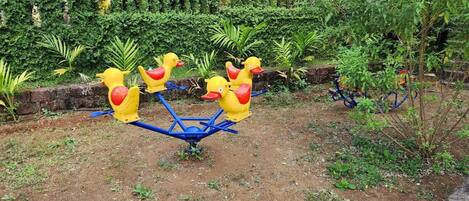 Un sube y baja con asientos con forma de pato, amarillo y rojo, en un jardín con abundante vegetación y un muro de piedra.