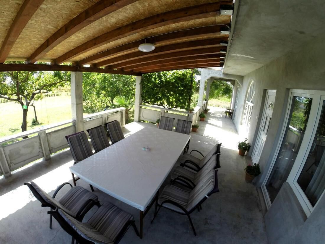 Un patio techado al aire libre con una mesa grande y sillas, rodeado de plantas en macetas y vistas a la vegetación.
