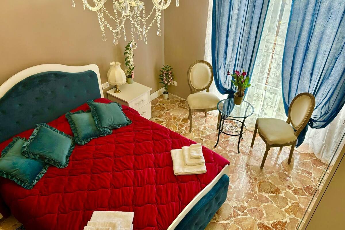 Ein Schlafzimmer mit roter Bettdecke, einem Kronleuchter, einem blauen Sessel und einem kleinen Tisch mit Pflanze.