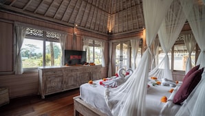 A bedroom with a wooden ceiling, a TV on a wooden dresser, a bed with white bedding and a canopy, and a view of the outdoors through large windows. - Uma Caming Villa Ubud (Payangan)