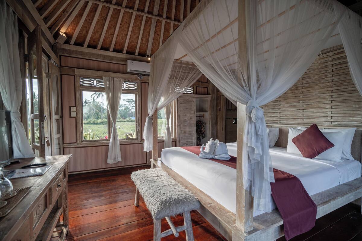 Une chambre avec un plancher en bois, un lit avec une literie blanche et bordeaux, un banc et un baldaquin.