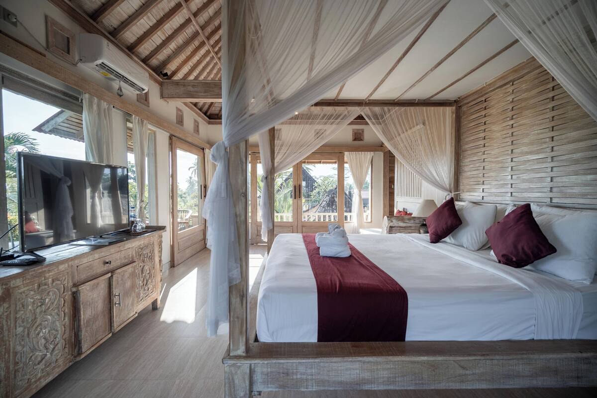 Une chambre avec un grand lit, une armoire en bois, une télévision et un balcon avec des rideaux.