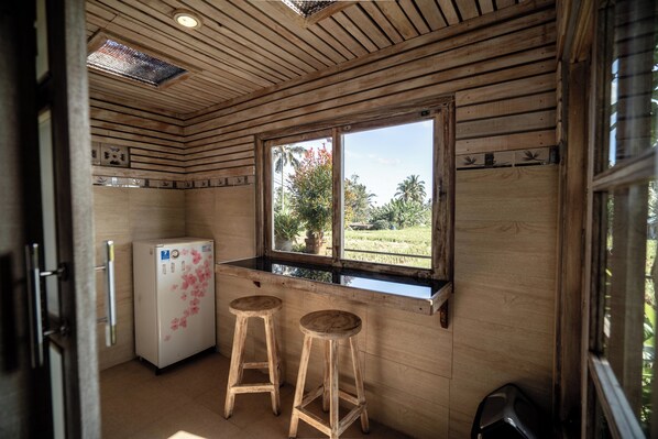 A compact kitchen with wooden walls, a refrigerator, two stools, and a window with a view of greenery. - Uma Caming Villa Ubud (Payangan)