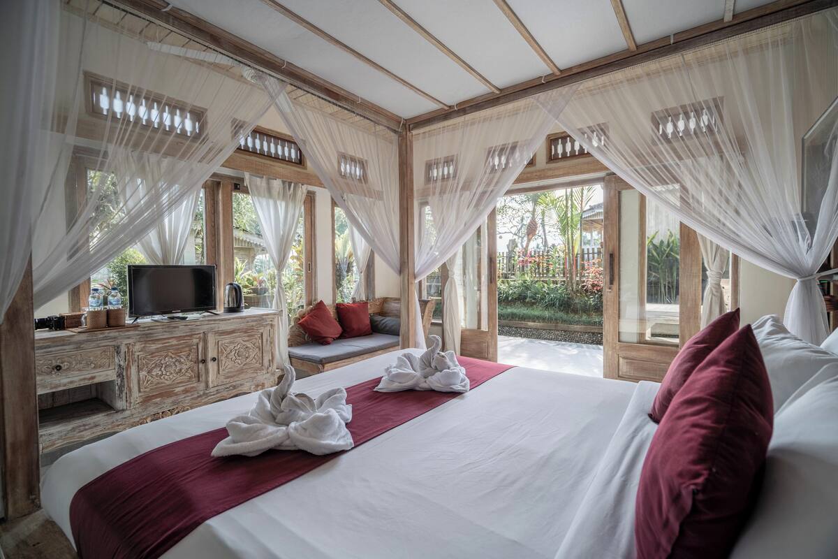 Une chambre spacieuse avec un grand lit, un téléviseur posé sur une commode en bois et une vue sur le jardin grâce à de grandes fenêtres.