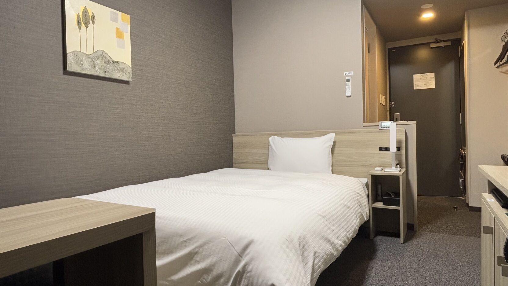 A hotel room with a bed, a desk, a chair, a wardrobe, a mirror, and a painting on the wall.