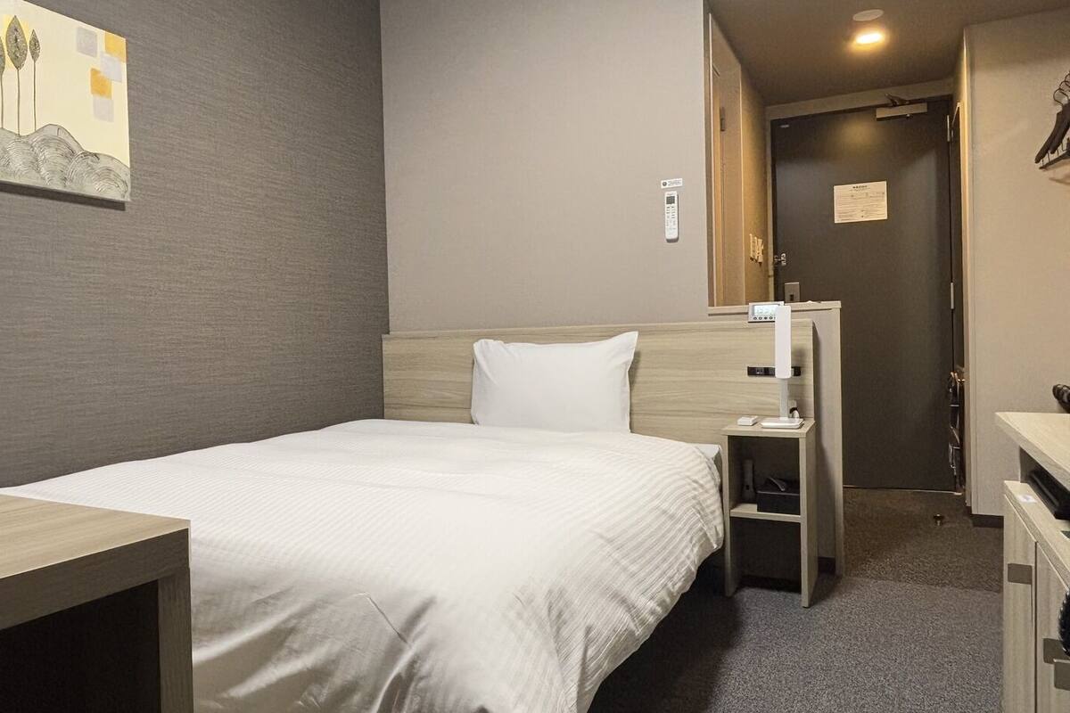 A hotel room with a bed, a desk, a chair, a wardrobe, a mirror, and a painting on the wall.