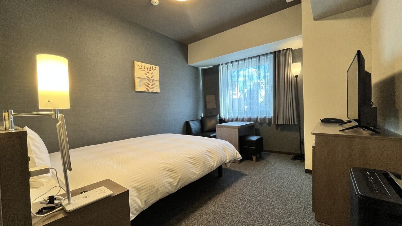 A hotel room with a bed, desk, chair, lamp, TV, and a small table.