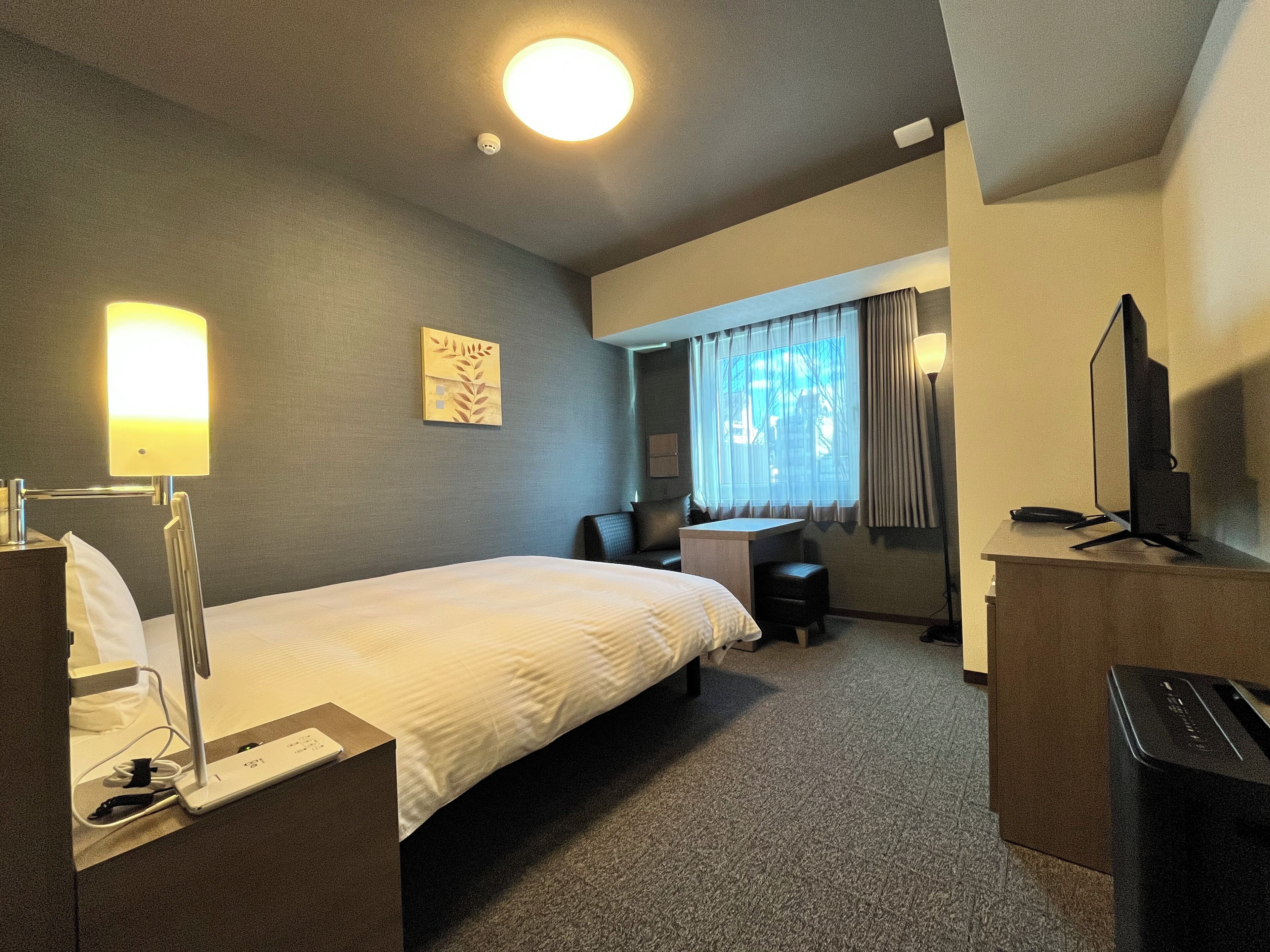 A hotel room with a bed, desk, chair, lamp, TV, and a window with curtains.