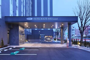 A szálloda bejárata egy fedett gyalogúton keresztül vezet, a területen parkolóhelyek találhatók, és egy tábla jelzi a „HOTEL ROUTE INN Grand” nevet.