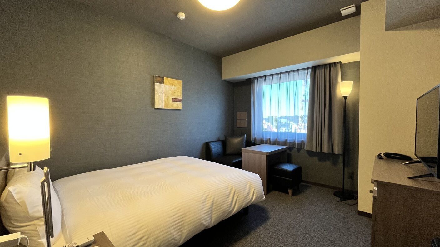 A hotel room with a bed, desk, chair, lamp, and a view of the city through the window.