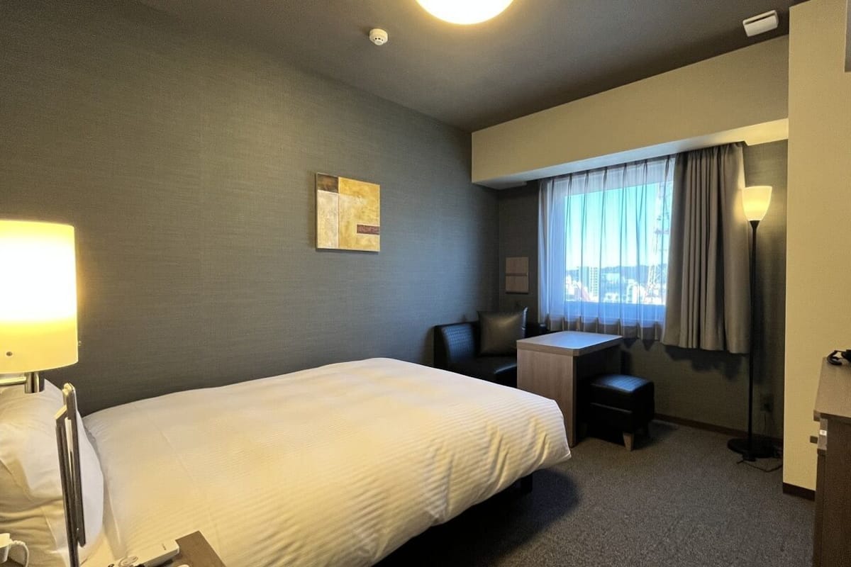 Une chambre d’hôtel équipée d’un lit, d’un bureau, d’une chaise, d’une lampe et offrant une vue sur la ville par la fenêtre.
