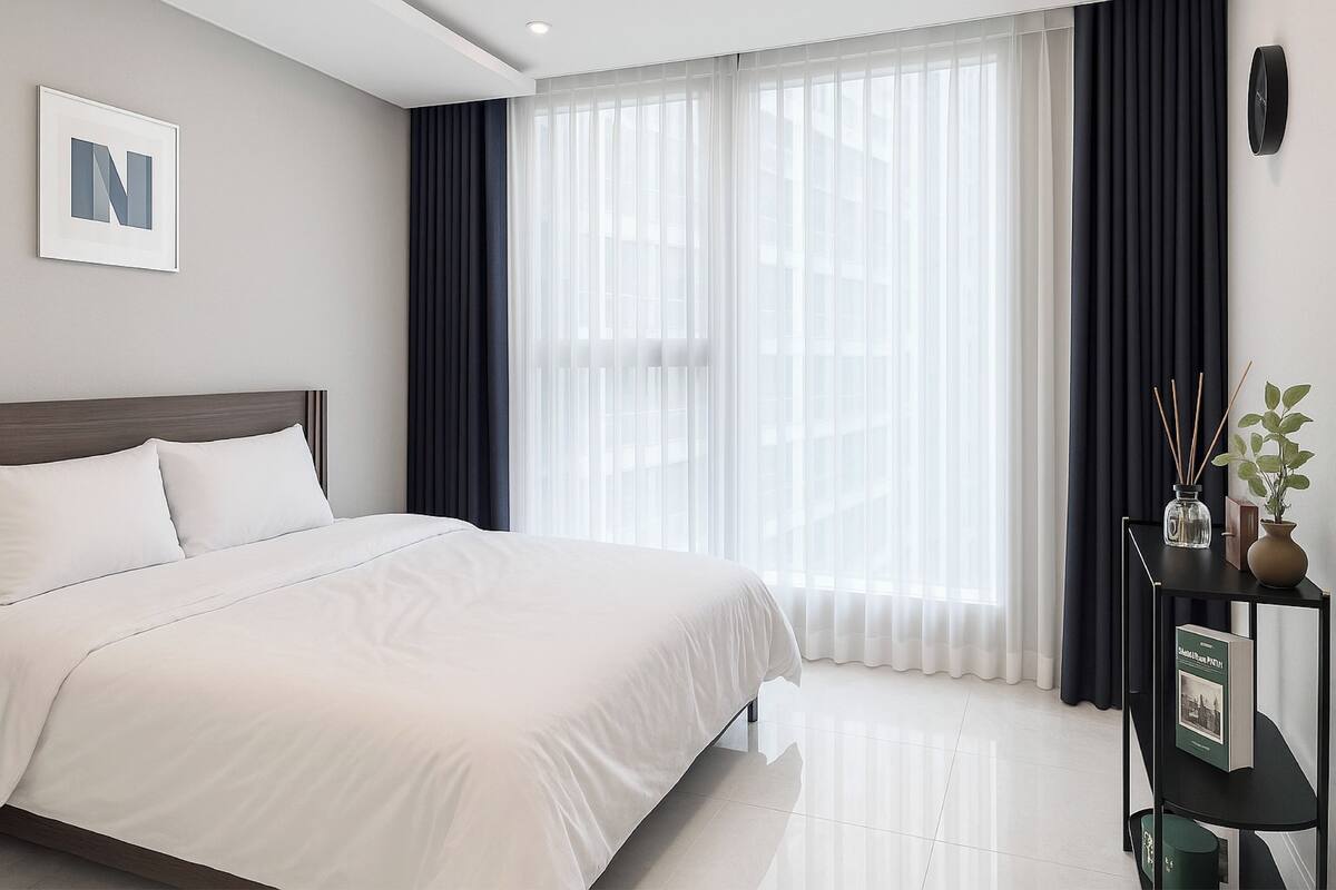 Une chambre d’hôtel moderne avec un grand lit, une table de chevet sur laquelle se trouvent un livre et un vase, et une vue sur la ville à travers de fines rideaux.