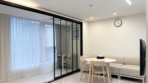 Een moderne woonkamer met een glazen wand, een ronde tafel met stoelen, een bank, een flatscreen televisie en een wandklok.