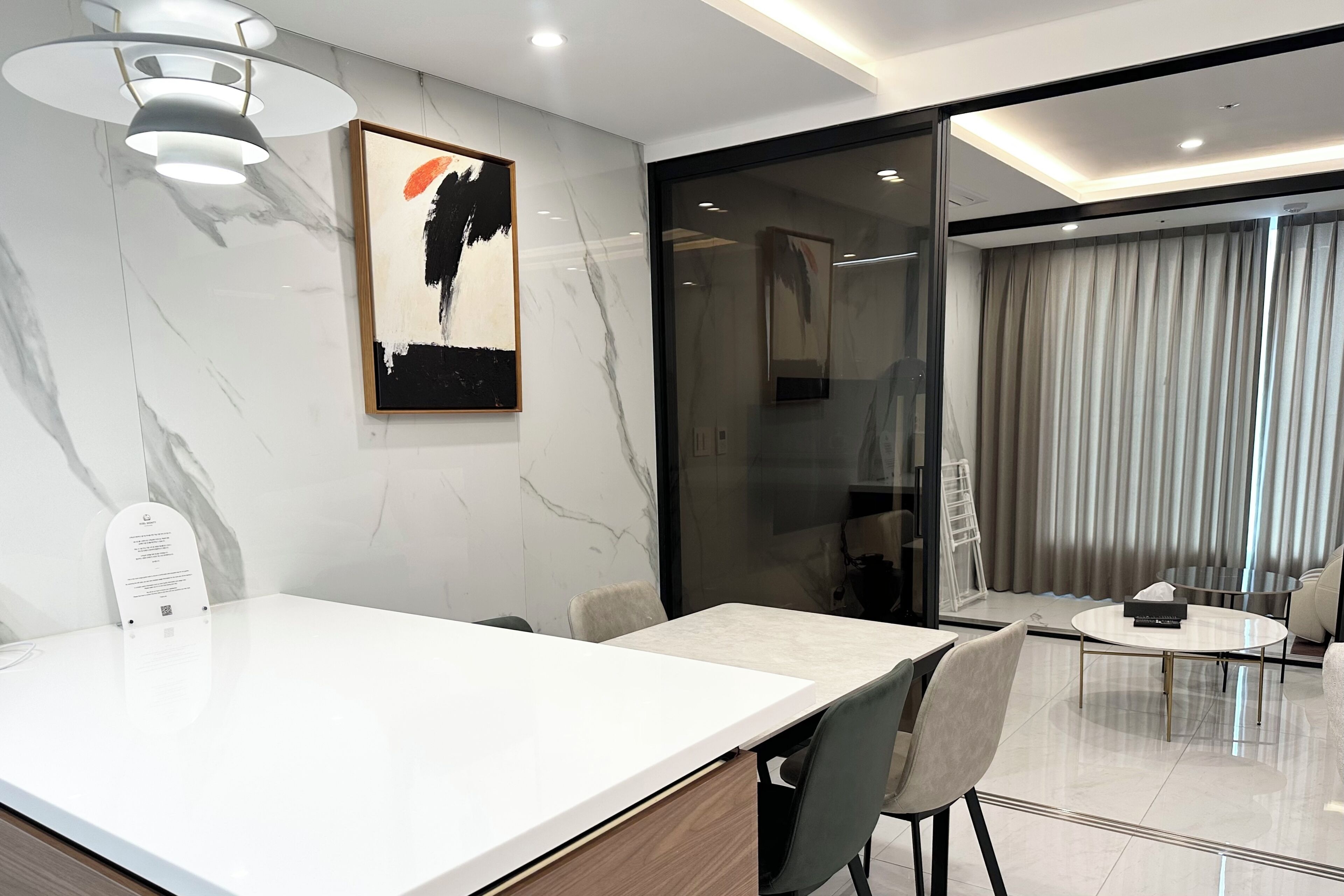 Een moderne hotelkamer met een marmeren wand, een zithoek met een witte tafel en stoelen, en een groot raam met gordijnen.