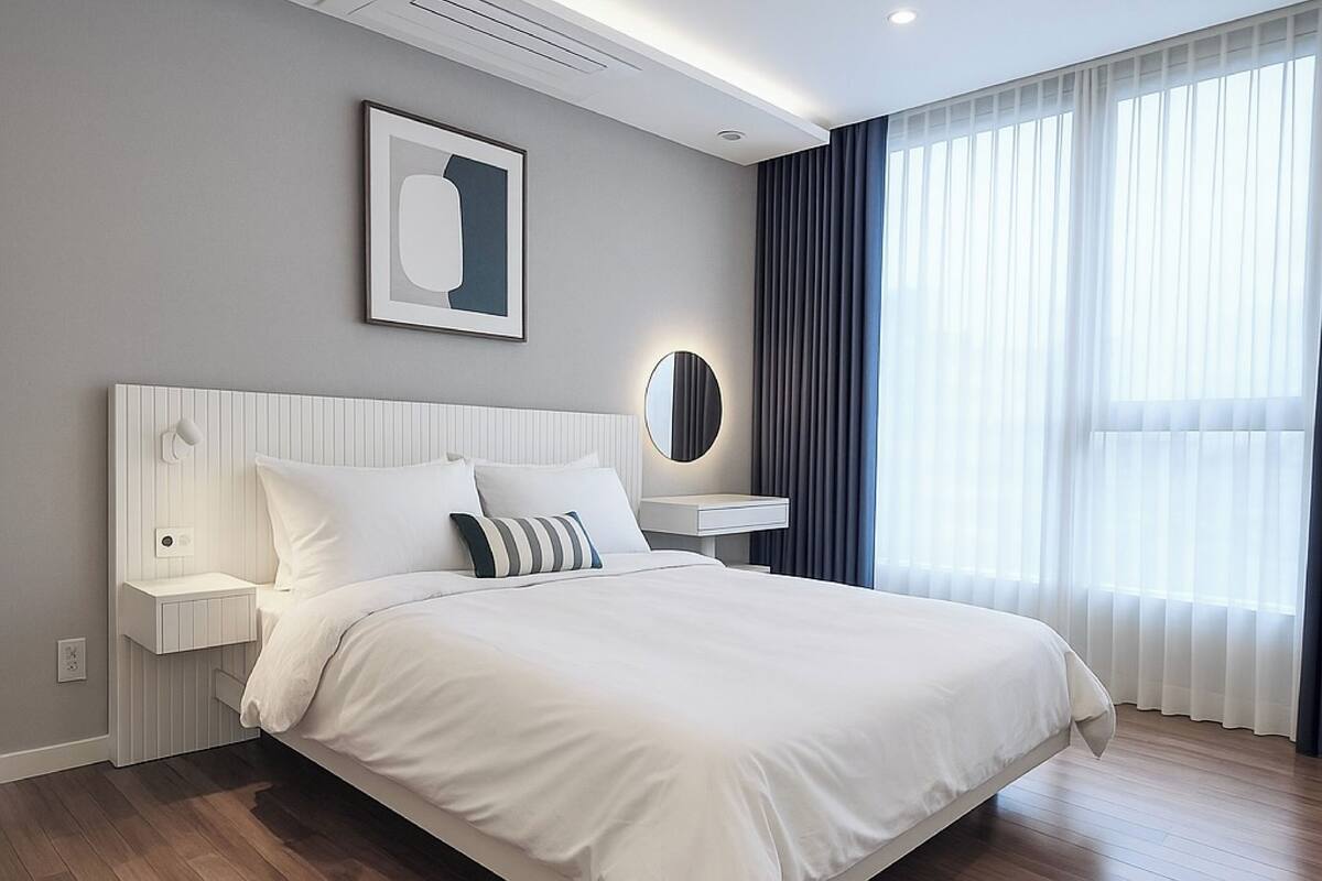 Une chambre d’hôtel moderne avec un grand lit, une table de chevet avec une lampe, un miroir fixé au mur et une grande fenêtre avec des rideaux.