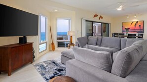 Un salon doté d’un grand canapé d’angle gris, d’un téléviseur à écran plat posé sur un meuble en bois, d’une guitare en bois et offrant une vue sur l’océan grâce à de grandes fenêtres.