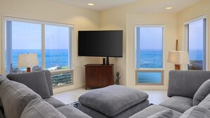 Un salon spacieux doté d’un grand téléviseur à écran plat fixé au mur, de canapés gris et offrant une vue sur l’océan grâce à de grandes fenêtres.