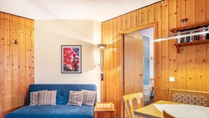 Um quarto com um sofá azul, uma mesa de apoio de madeira, um pequeno banquinho de madeira, uma mesa de jantar de madeira com cadeiras e uma estante de livros de madeira.
