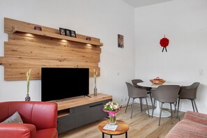 Ein modernes Wohnzimmer mit Flachbildfernseher, Holzvertäfelung, einem roten Sofa, einem kleinen runden Tisch mit einer Vase und einem Essbereich mit weißem Tisch und grauen Stühlen.