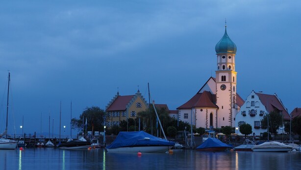 A waterfront with a prominent church featuring a clock tower and a spire, surrounded by calm water and boats. - Apartment 'Denn' with Shared Garden, Balcony and Wi-Fi (Langenargen)