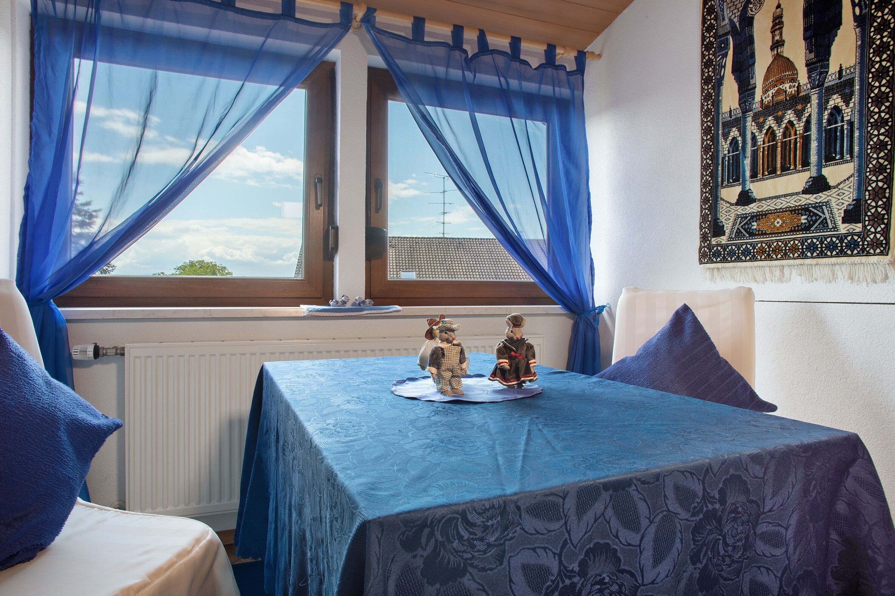 Une pièce avec une table dressée pour deux personnes, une fenêtre ornée de rideaux bleus, un radiateur et une tapisserie accrochée au mur.