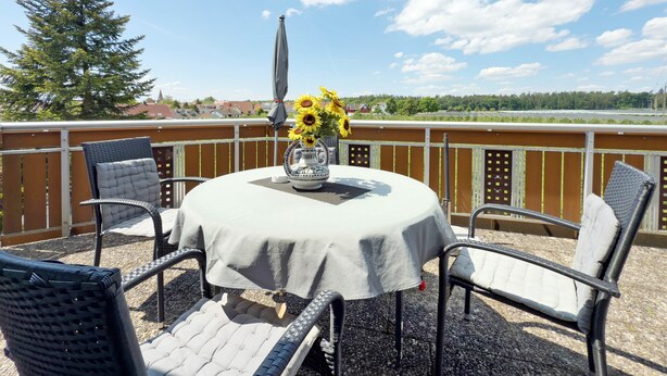 Una mesa redonda cubierta con un mantel blanco, rodeada por cuatro sillas con asientos y respaldos acolchados. Un jarrón con girasoles está sobre la mesa.