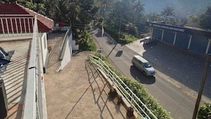 A view from a balcony overlooking a road with a white car, a cocoa chocolate factory, and lush greenery in the background. - Aloha Homestay Munnar (Anachal)