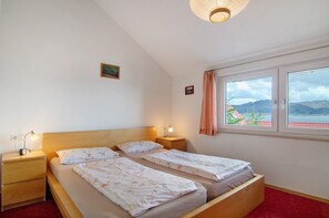 A room with two beds, a wooden headboard, bedside lamps, a small table, and a window with a view of hills.