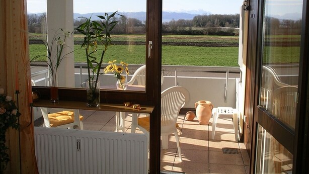 Ein Balkon mit Blick auf ein grünes Feld und Berge, weiße Stühle, ein Tisch mit Blumen in einer Vase und ein Heizkörper.