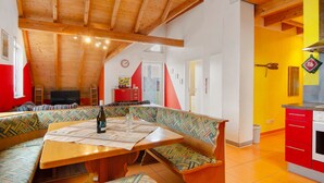 A cozy room with a wooden ceiling, a dining table with a bottle and glass, a colorful bench, and a kitchen area with red cabinets. - Saupp Apartment with Garden and Wi-Fi (Immenstaad am Bodensee)