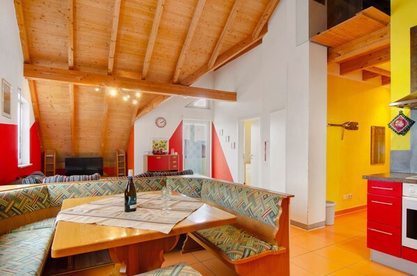 A cozy room with a wooden ceiling, a dining table with a bottle and glass, a colorful bench, and a kitchen area with red cabinets.