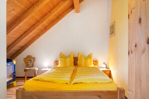 A room with a sloped ceiling, a bed with yellow bedding, a wooden chair, a bedside table with a lamp, and a framed picture on the wall.