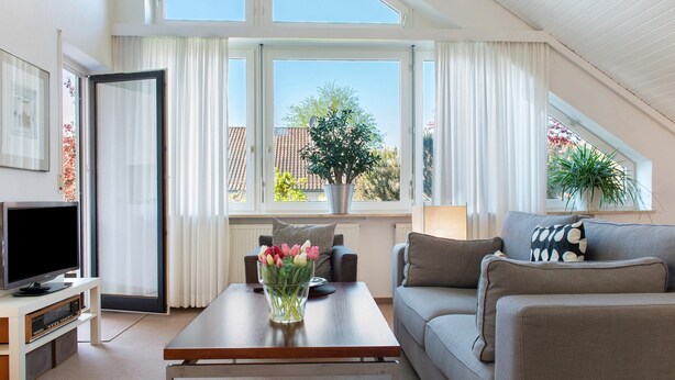 Een moderne woonkamer met een flatscreen televisie, een salontafel met een bloevas, een bank met gekleurde kussens en grote ramen met gordijnen.
